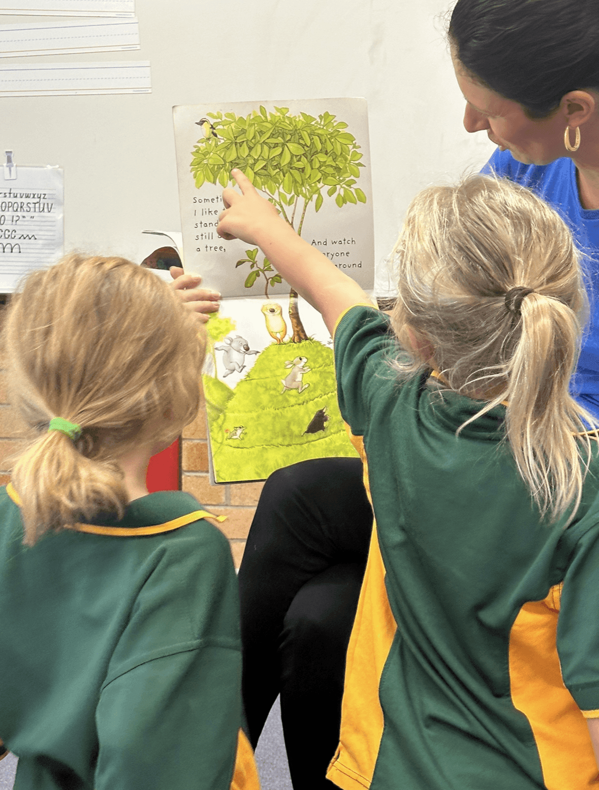 Two children sitting with a teacher, pointing at an illustrated book page featuring a tree and animals.