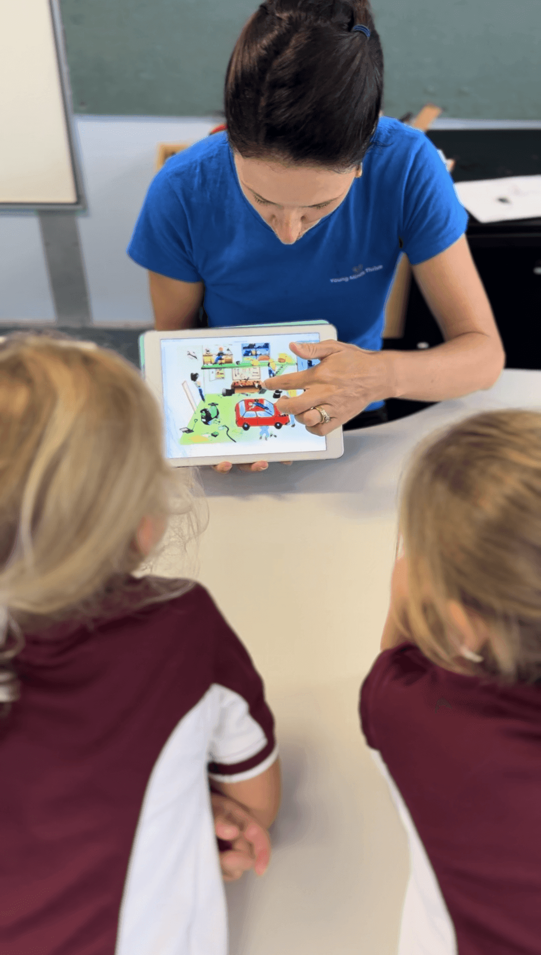 Individual showing a tablet displaying a colorful illustration to two children in maroon and white shirts.