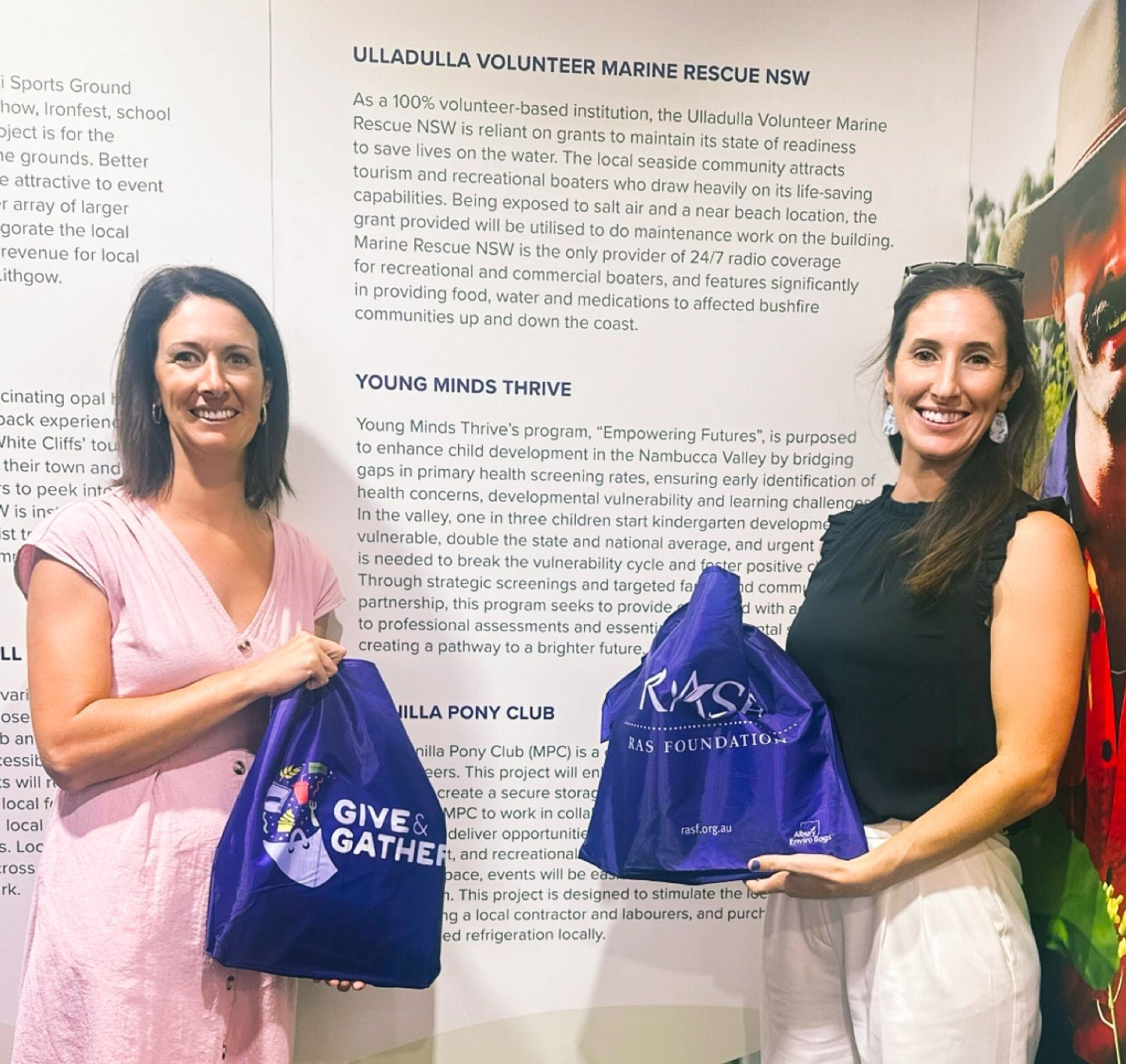 Two women smiling and holding purple bags in front of informational posters about community programs.